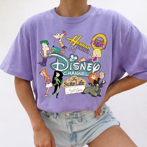 May include: A lilac t-shirt with a Disney Channel graphic. The design features cartoon characters from shows like Phineas and Ferb, Hannah Montana, and Kim Possible. The Disney logo and show titles are also included.