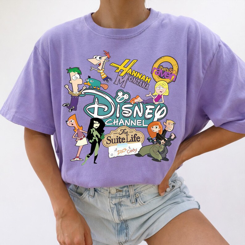 May include: A lilac t-shirt with a Disney Channel graphic. The design features cartoon characters from shows like Phineas and Ferb, Hannah Montana, and Kim Possible. The Disney logo and show titles are also included.