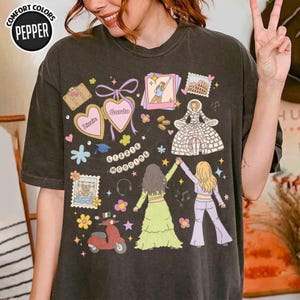 May include: A charcoal gray short-sleeve t-shirt featuring a graphic print with the text Lizzie McGuire. The design includes illustrations of a scooter, heart-shaped lockets, a dress, and two figures, accented with colorful flowers and stars.