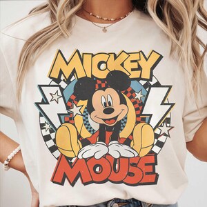 K&ouml;nnte beinhalten: Cremefarbenes T-Shirt mit einem Retro-Grafikdesign von Mickey Mouse. Das Design enth&auml;lt die Worte "Mickey Mouse" in fetter, farbenfroher Schrift, mit Mickey in der Mitte. Das Shirt hat eine klassische, Vintage-&Auml;sthetik.