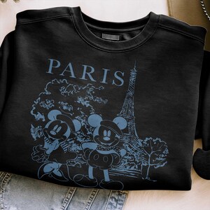 K&ouml;nnte beinhalten: Schwarzes Sweatshirt mit Rundhalsausschnitt und blauem Aufdruck von Mickey und Minnie Mouse vor dem Eiffelturm in Paris. Das Wort "PARIS" ist blau &uuml;ber dem Aufdruck gedruckt.