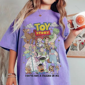 May include: A purple t-shirt featuring a graphic of characters from the animated film "Toy Story." The design includes the film's title and the phrase "You've Got a Friend in Me." The shirt has a relaxed fit.