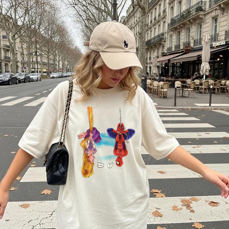 May include: Cream-colored t-shirt featuring a cartoon illustration of Rapunzel and Spider-Man hanging upside down. The shirt is paired with a beige baseball cap with a dark blue logo. A black shoulder bag with a chain strap completes the look.