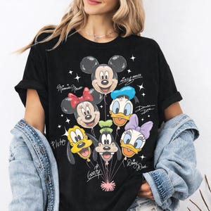 May include: A black short-sleeve t-shirt featuring a graphic print of Mickey Mouse, Minnie Mouse, Donald Duck, Pluto, Goofy, and Daisy Duck as balloons. The names of each character are written in white script next to their respective balloon faces.