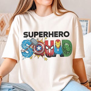 May include: Cream-colored t-shirt with the words "SUPERHERO SQUAD" in black block letters. The word "SQUAD" is designed with superhero-themed graphics, including Thor's hammer, Captain America's shield, and Iron Man's helmet.