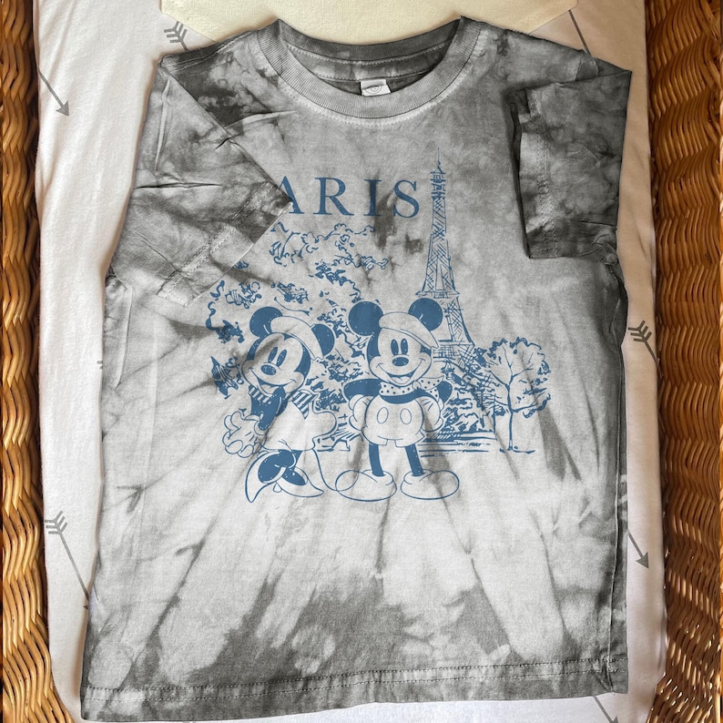 K&ouml;nnte beinhalten: Graues T-Shirt mit Batikmuster und einem blauen Aufdruck von Mickey und Minnie Mouse vor dem Eiffelturm und dem Wort "PARIS". Das T-Shirt liegt gefaltet auf einer wei&szlig;en Oberfl&auml;che mit grauen Pfeilen.