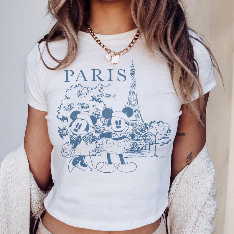 K&ouml;nnte beinhalten: Cremefarbenes T-Shirt mit einem blauen Aufdruck von Mickey und Minnie Mouse vor dem Eiffelturm in Paris. Das Wort "PARIS" ist &uuml;ber dem Aufdruck gedruckt. Das T-Shirt hat kurze &Auml;rmel und einen Rundhalsausschnitt.
