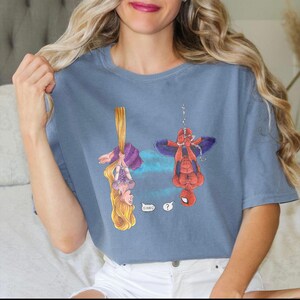 May include: A blue t-shirt featuring cartoon illustrations of Rapunzel and Spider-Man hanging upside down. Rapunzel is holding her long blonde hair, and Spider-Man is suspended by a web. Speech bubbles include the words "Oh Hai!" and a question mark.
