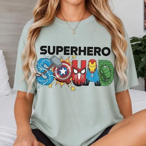 May include: A light green t-shirt with the words "SUPERHERO SQUAD" in black and colorful superhero-themed graphics. The "S" features Thor's hammer and lightning, the "U" has Captain America's shield, and the "D" has the Hulk's fist.