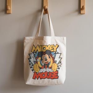 K&ouml;nnte beinhalten: Eine cremefarbene Tragetasche mit einer farbenfrohen Mickey Mouse-Grafik. Das Design beinhaltet die Worte "Mickey Mouse" in einer Retro-Schriftart, mit Blitzen und Sternen. Die Tasche hat lange Griffe zum Tragen.