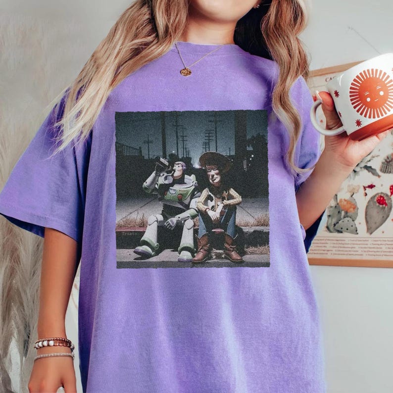 May include: A lilac-colored t-shirt featuring a graphic print of Buzz Lightyear and Woody from Toy Story, sitting together. The image has a vintage, slightly faded aesthetic. The shirt is a relaxed fit.