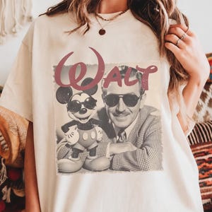 K&ouml;nnte beinhalten: Ein cremefarbenes T-Shirt mit einem Grafikdruck von Walt Disney und Mickey Mouse, die beide eine Sonnenbrille tragen. Das Wort "WALT" ist in einer gro&szlig;en, stilisierten kastanienbraunen Schrift &uuml;ber dem schwarz-wei&szlig;en Foto des Paares abgedruckt.