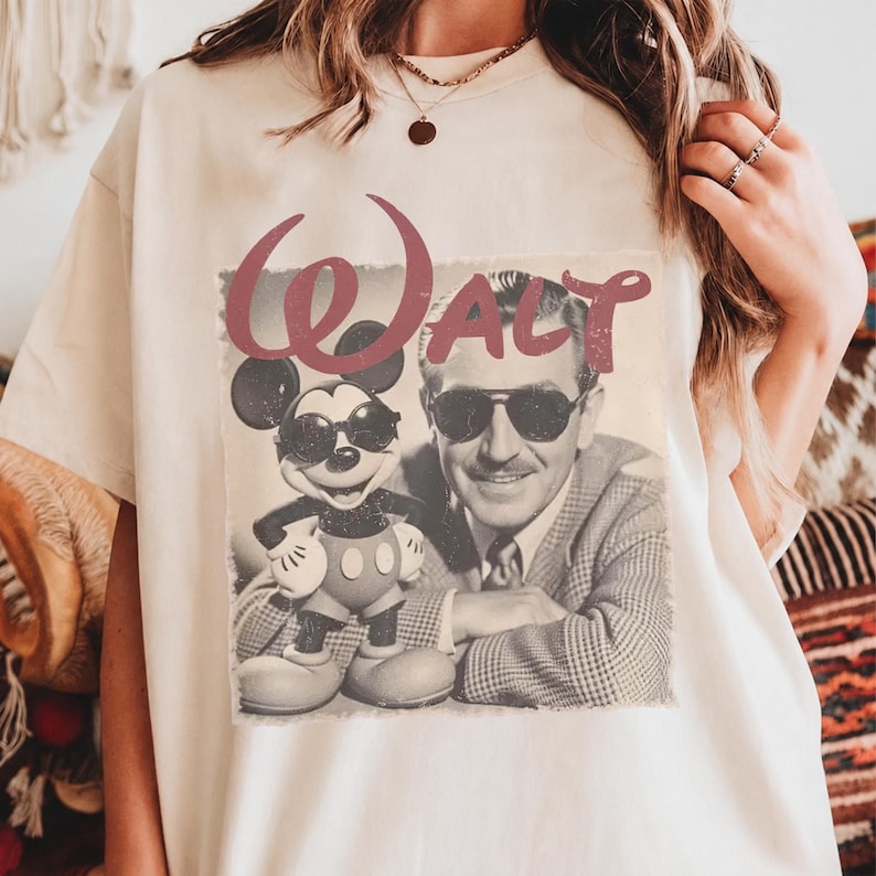 K&ouml;nnte beinhalten: Ein cremefarbenes T-Shirt mit einem Grafikdruck von Walt Disney und Mickey Mouse, die beide eine Sonnenbrille tragen. Das Wort "WALT" ist in einer gro&szlig;en, stilisierten kastanienbraunen Schrift &uuml;ber dem schwarz-wei&szlig;en Foto des Paares abgedruckt.