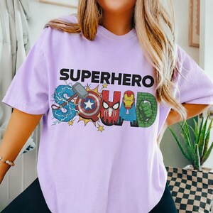 May include: A light purple t-shirt with the words "SUPERHERO SQUAD" in black block letters. The word "SQUAD" is designed with superhero logos and graphics, including Thor's hammer, Captain America's shield, and Iron Man's mask.