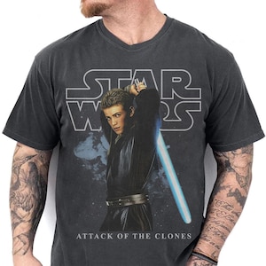 May include: Dark gray v-neck t-shirt featuring a graphic of a character from Star Wars: Attack of the Clones holding a blue lightsaber. The Star Wars logo is above the graphic.