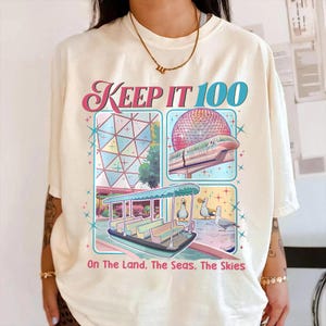 May include: Cream-colored t-shirt featuring a colorful graphic with the text "KEEP IT 100." The graphic includes images of a geometric structure, a monorail, a boat, and seagulls. The phrase "On The Land. The Seas. The Skies" is also printed on the shirt.