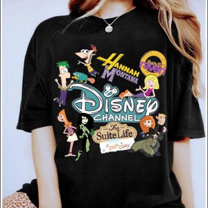 May include: Black t-shirt with a vibrant graphic showcasing Disney Channel characters such as Phineas and Ferb, Hannah Montana, Kim Possible, and The Suite Life of Zack & Cody. The Disney Channel logo is visible.