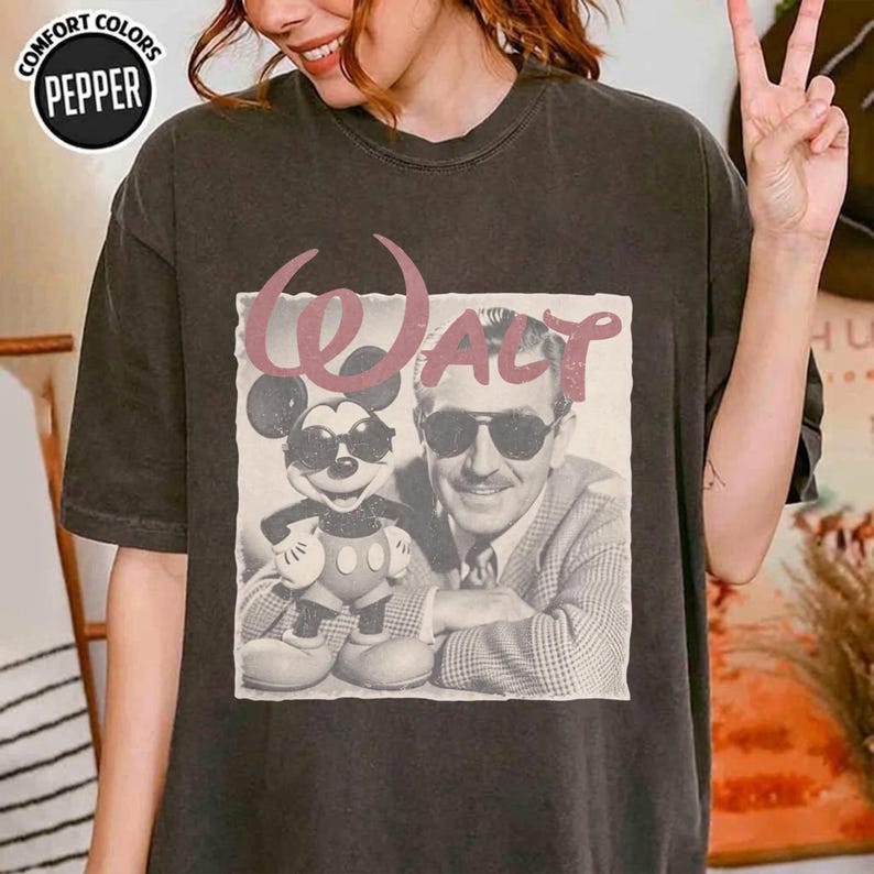 K&ouml;nnte beinhalten: Ein anthrazitfarbenes Kurzarm-T-Shirt mit einem Grafikdruck von Walt Disney und Mickey Mouse, die beide eine Sonnenbrille tragen. Der Text Walt ist in einer gro&szlig;en, stilisierten rosa Schrift &uuml;ber den Figuren gedruckt. Comfort Colors Etikett.