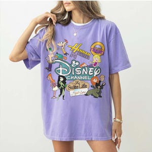 May include: A lilac t-shirt with a Disney Channel graphic. The design features cartoon characters and logos from shows such as "Hannah Montana" and "The Suite Life of Zack & Cody". The shirt has short sleeves and a crew neck.