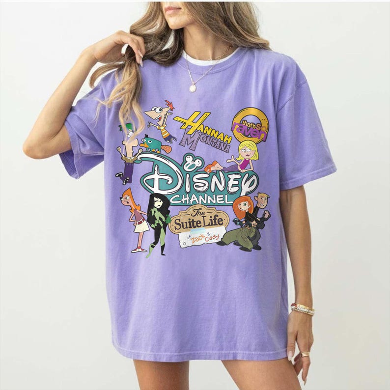 May include: A lilac t-shirt with a Disney Channel graphic. The design features cartoon characters and logos from shows such as "Hannah Montana" and "The Suite Life of Zack & Cody". The shirt has short sleeves and a crew neck.