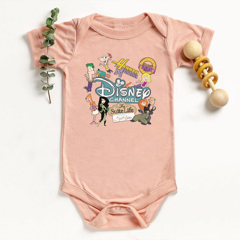May include: A pale pink baby bodysuit with a Disney Channel graphic. The graphic features characters from shows like Phineas and Ferb, Hannah Montana, and Kim Possible. A wooden rattle is positioned to the right of the bodysuit.