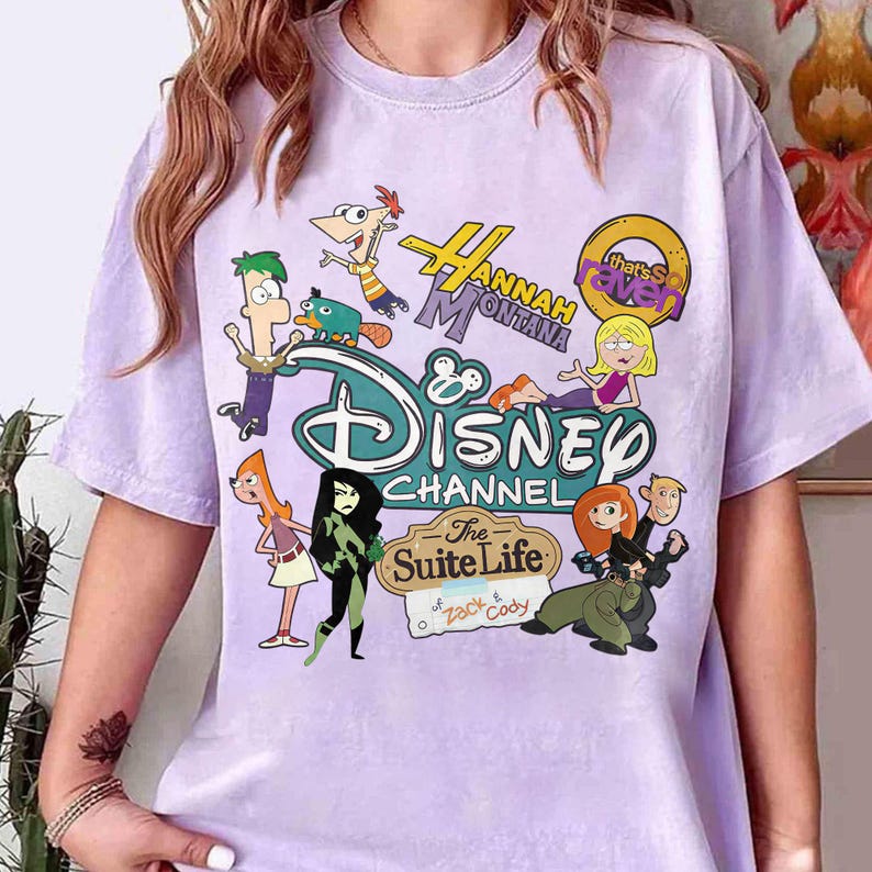 May include: A lilac t-shirt with a Disney Channel design. The graphic includes cartoon characters from shows like Hannah Montana, Kim Possible, and The Suite Life of Zack & Cody. The top has a crew neck and short sleeves.
