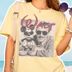 K&ouml;nnte beinhalten: Ein hellgelbes Kurzarm-T-Shirt mit einem schwarz-wei&szlig;en Grafikdruck von Walt Disney und Mickey Mouse, die beide eine Sonnenbrille tragen. Das Wort Walt ist in einer gro&szlig;en, verwaschenen kastanienbraunen Schreibschrift dar&uuml;ber gedruckt.