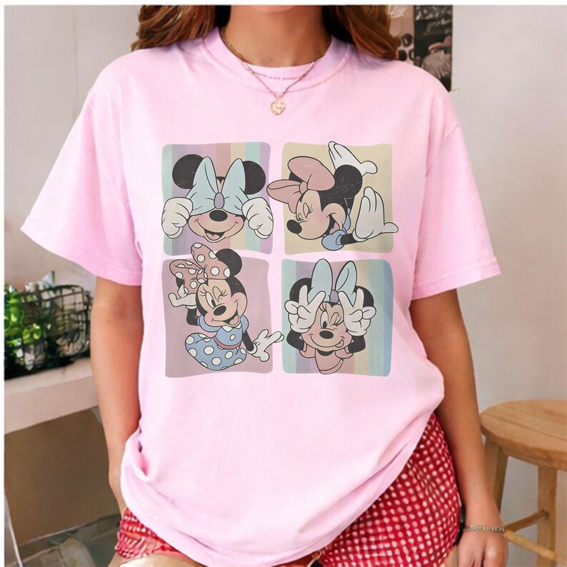 May include: A light pink t-shirt featuring a four-panel graphic of Minnie Mouse in various poses. Each panel has a pastel background. Minnie is wearing a bow and a dress in the bottom left panel. The shirt has short sleeves.