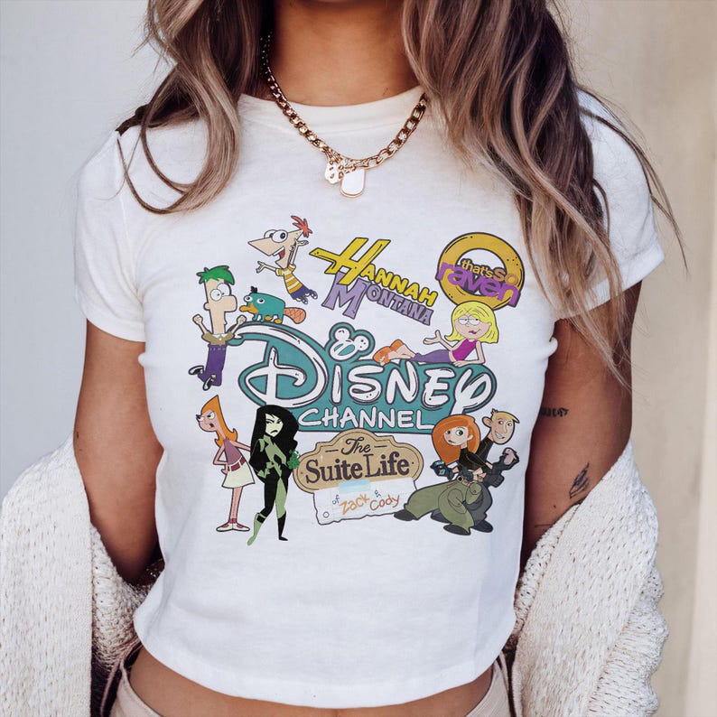May include: A white t-shirt with a vibrant graphic showcasing Disney Channel characters and logos, such as "Hannah Montana" and "The Suite Life of Zack & Cody." The design incorporates cartoon illustrations of several characters. The shirt has short sleeves.