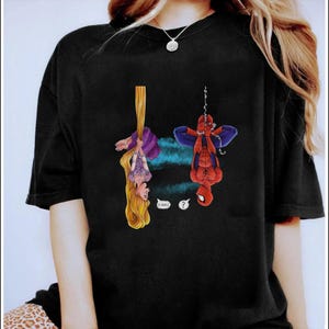 May include: Black t-shirt featuring a cartoon illustration of Rapunzel hanging from her hair and Spider-Man hanging upside down. Rapunzel says "O HAI!" and Spider-Man has a question mark above his head. The shirt has a crew neck.