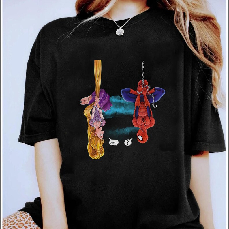May include: Black t-shirt featuring a cartoon illustration of Rapunzel hanging from her hair and Spider-Man hanging upside down. Rapunzel says "O HAI!" and Spider-Man has a question mark above his head. The shirt has a crew neck.