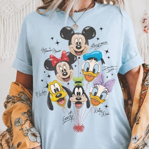 May include: A light blue t-shirt featuring a graphic print of Mickey Mouse, Minnie Mouse, Donald Duck, Pluto, Goofy, and Daisy Duck as balloons. Each character is labeled with their name in black script, surrounded by small black star icons.