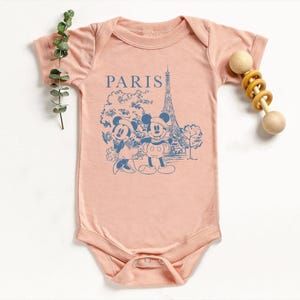 K&ouml;nnte beinhalten: Hellrosa Baby-Body mit blauem Aufdruck von Mickey und Minnie Mouse vor dem Eiffelturm, mit dem Wort "PARIS" dar&uuml;ber. Auf der rechten Seite des Bodys befindet sich eine Holzrassel.