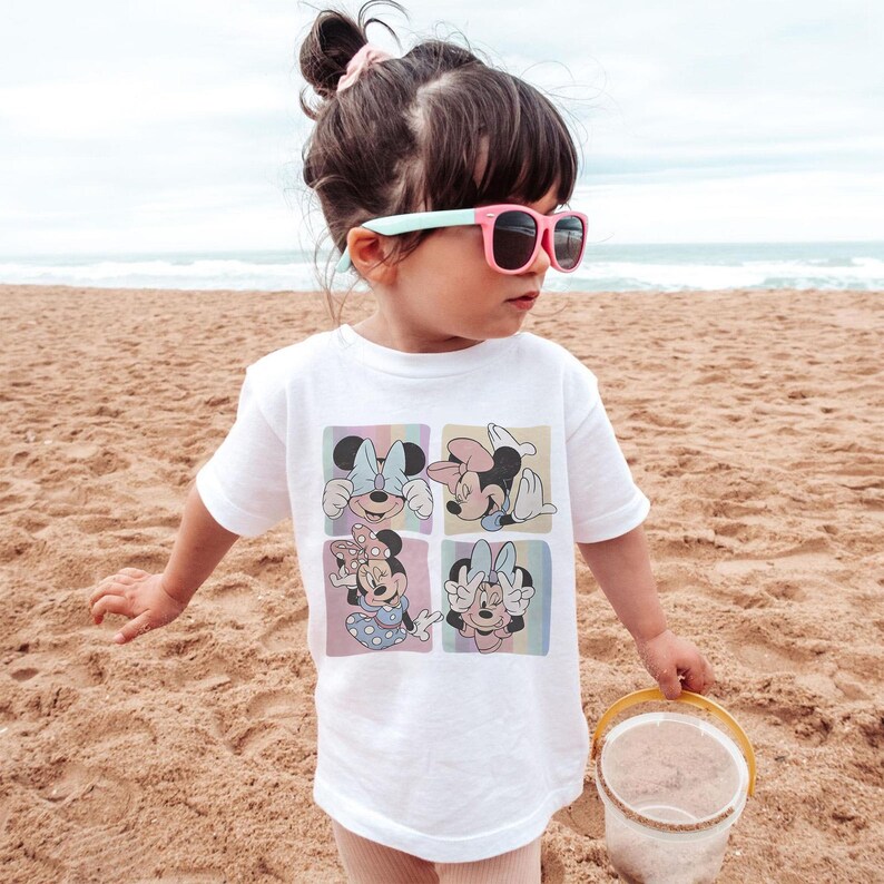May include: White t-shirt featuring four squares with Minnie Mouse illustrations in pastel colors. The child is wearing pink sunglasses and is standing on a sandy beach. A small yellow bucket is in the child's hand.