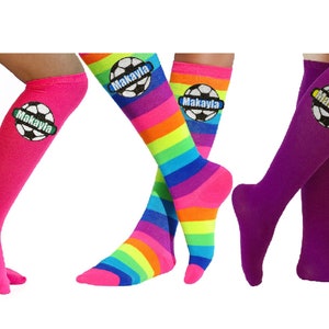 May include: Three pairs of knee-high socks with a soccer ball design and the name "Makayla" on each. One pair is pink, one is rainbow, and one is purple.