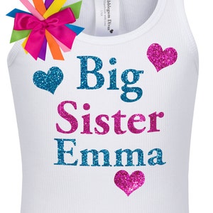 May include: White tank top with the text "Big Sister Emma" in blue and pink glitter lettering. The shirt has three glitter hearts, one blue and two pink.