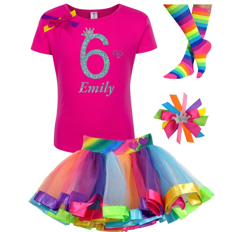 May include: A pink t-shirt with a glittery teal number 6 and a crown, the name "Emily" is written below the number. A rainbow tulle skirt with a rainbow stripe waistband and a glittery pink heart. A rainbow striped pair of socks and a rainbow hair bow with a silver crown.