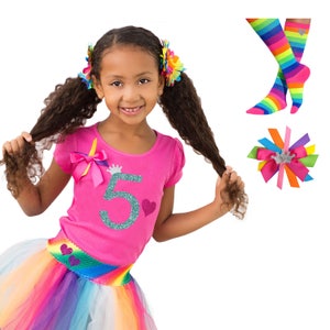 May include: A girl wearing a pink shirt with a glittery number 5 and a crown on it. She is also wearing a rainbow tutu and has rainbow hair bows. The image also includes a pair of rainbow socks and a rainbow hair bow with a crown.