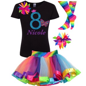 以下が含まれることがあります： ターコイズブルーの数字8、紫色の蝶、そして「Nicole」という名前が紫色のグリッターで書かれた黒いTシャツ。紫、ピンク、黄色、緑、青、オレンジのリボンが付いたレインボーチュチュ。レインボーストライプの靴下と、紫色の蝶が付いたレインボーリボン。