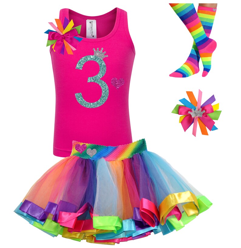 May include: A pink tank top with a glittery teal number "3" and a crown on it. The tank top is paired with a rainbow tulle skirt with colorful satin ribbon trim. A rainbow striped knee-high sock and a rainbow hair bow with a crown are also included.