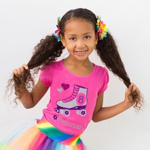 May include: A young girl wearing a pink shirt with a silver roller skate graphic and the number 8. The shirt also has the word "princess" on it. She is wearing a rainbow tutu skirt and has her hair in pigtails with colorful flower clips.