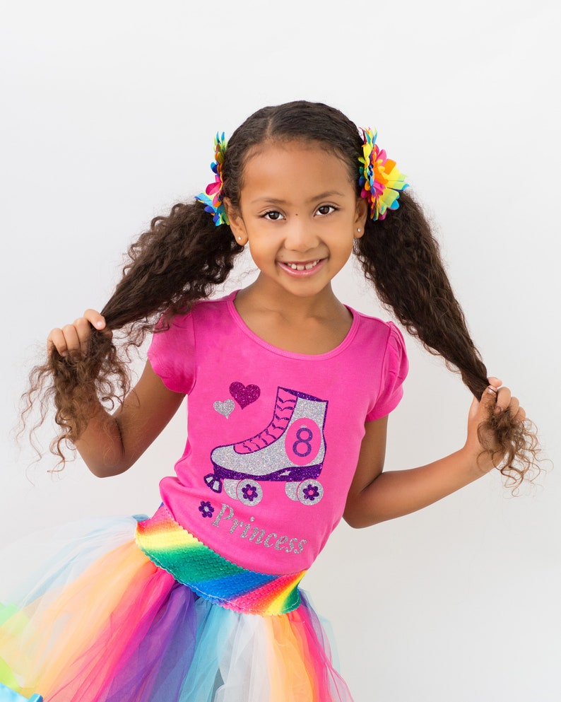 May include: A young girl wearing a pink shirt with a silver roller skate graphic and the number 8. The shirt also has the word "princess" on it. She is wearing a rainbow tutu skirt and has her hair in pigtails with colorful flower clips.