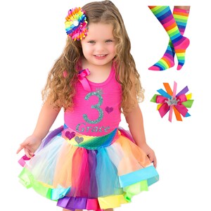 May include: A girl wearing a pink tank top with a glittery number 3 and the name "Grace" on it. She is also wearing a rainbow tutu skirt with a sequined mermaid tail. She has a rainbow flower hair clip in her hair. The image also includes a pair of rainbow striped socks and a rainbow bow hair clip.