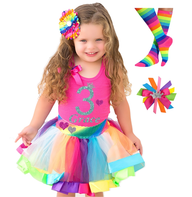 May include: A girl wearing a pink tank top with a glittery number 3 and the name "Grace" on it. She is also wearing a rainbow tutu skirt with a sequined mermaid tail. She has a rainbow flower hair clip in her hair. The image also includes a pair of rainbow striped socks and a rainbow bow hair clip.