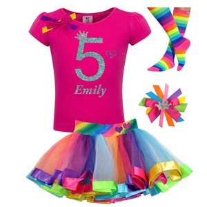 May include: A pink t-shirt with a glittery turquoise number 5 and a crown, the name "Emily" is written below the number. A rainbow tulle skirt with a rainbow stripe waistband and a matching rainbow striped bow with a crown. A pair of rainbow striped socks.