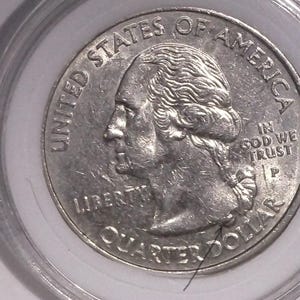 Puede incluir: Una moneda de un cuarto de dólar de los Estados Unidos de color plateado, encerrada en una cápsula protectora de plástico transparente. El anverso muestra el perfil de George Washington, el texto UNITED STATES OF AMERICA, IN GOD WE TRUST, LIBERTY y una marca de ceca P.