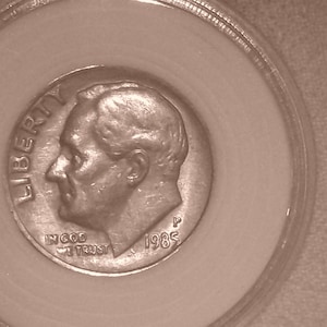 Könnte beinhalten: Eine 1985 P US-amerikanische Zehn-Cent-Münze in einer runden, durchsichtigen Kunststoff-Schutzhülle. Die silberfarbene Münze zeigt das nach links gewandte Profil von Franklin D. Roosevelt mit dem Text LIBERTY, IN GOD WE TRUST und dem Jahr 1985.