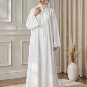 May include: A long, white dress with long sleeves and a button-down front. The dress features delicate lace trim on the cuffs and hem. The person in the image is wearing a matching head covering. The background includes a white wall and a beige curtain.