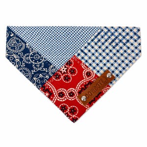 May include: A patchwork dog bandana featuring navy paisley, blue gingham, and red paisley patterns. A brown leather tag with the word "COWBOY" is attached with silver rivets. The bandana is designed to be worn around a dog's neck.