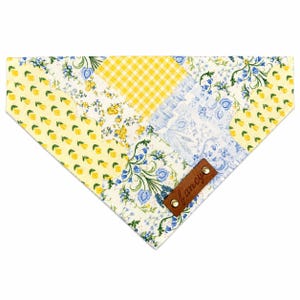 Peut inclure: Un bandana triangulaire pour animal avec un motif patchwork composé de vichy jaune, de fleurs jaunes et d'imprimés botaniques bleus et blancs. Une étiquette en similicuir marron avec le mot fancy en cursive est fixée par deux rivets.